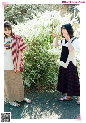 Two young women standing next to each other in front of bushes.